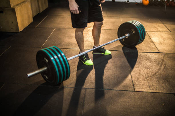 Weightlifter in neon green shoes standing behind a loaded barbell on a gym floor in warm sunlight