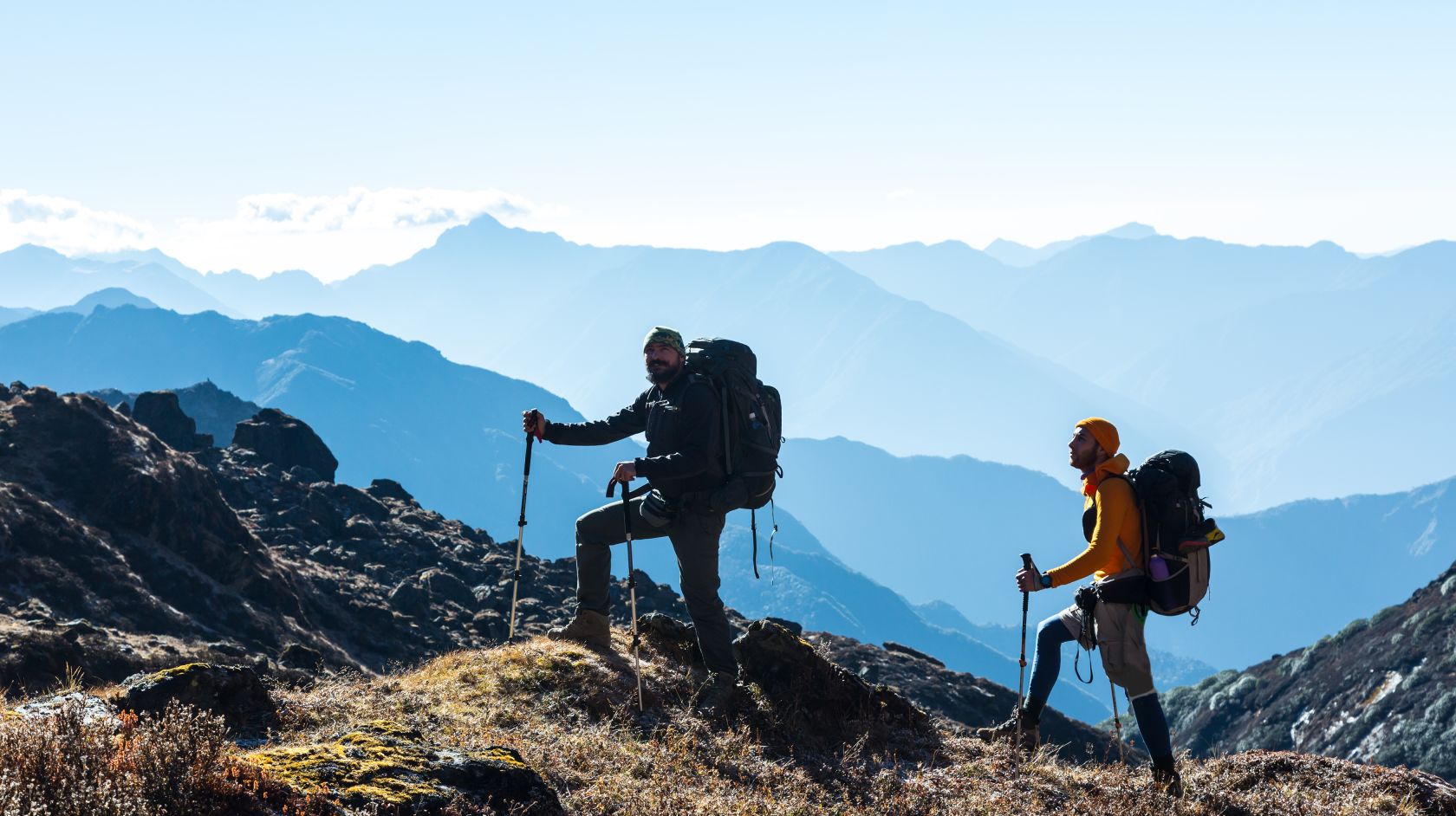 Two hikers with trekking poles and large backpacks crossing a rocky mountain ridge with layered blue peaks in the distance
